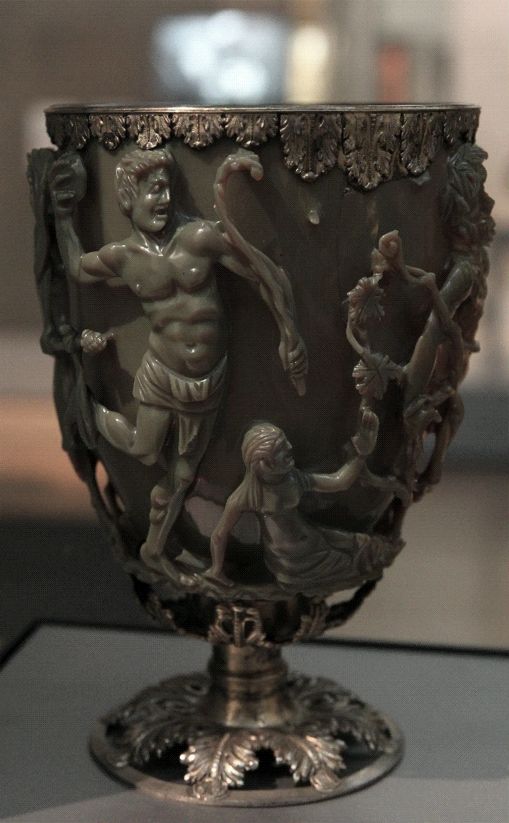 A Roman drinking cup of olive-green glass carved in deep openwork relief with struggling figures and vine tendrils, mounted with a gilt-metal rim and foot, standing in a museum case.