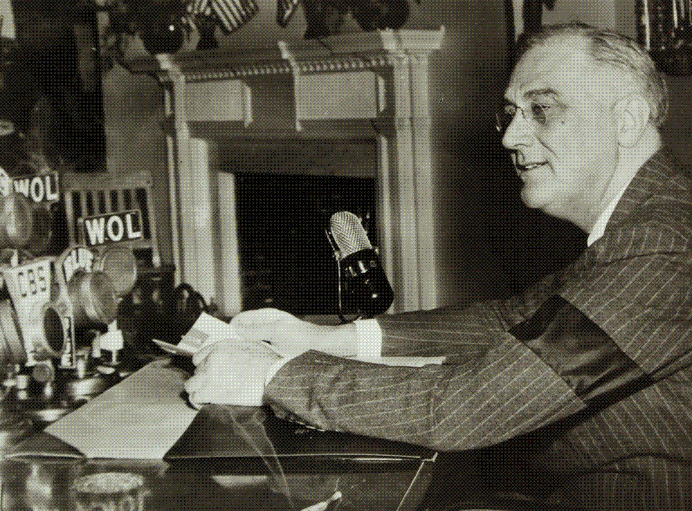 Franklin D. Roosevelt seated at a White House desk crowded with radio network microphones, papers in hand, delivering his broadcast address of 28 April 1942.