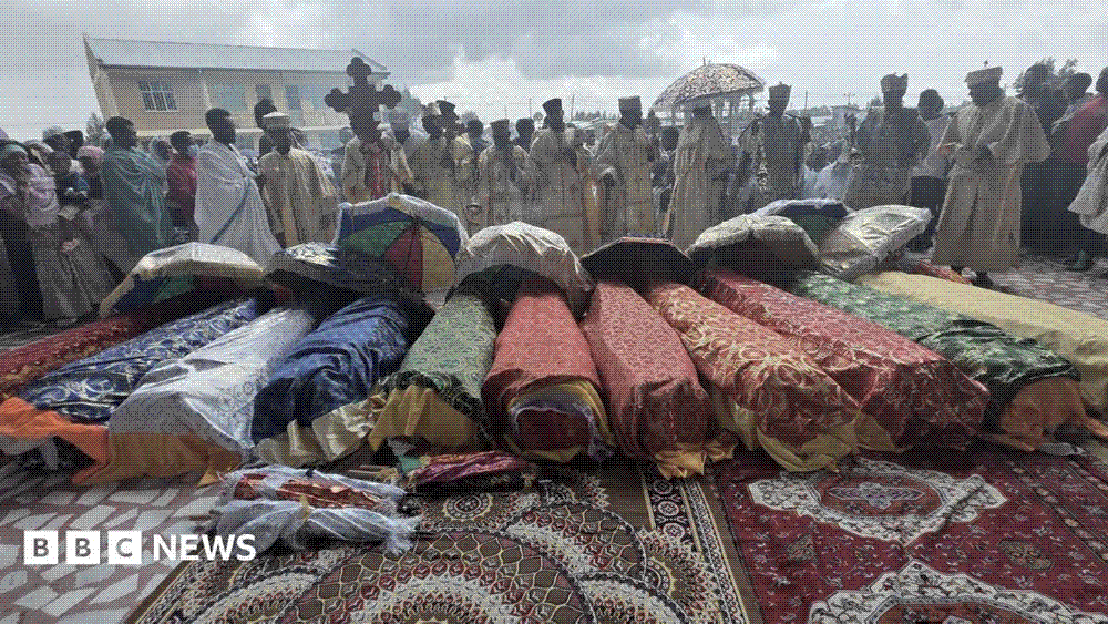Bodies wrapped in brightly patterned cloth laid in a row on carpets outside an Ethiopian Orthodox church while robed clergy and mourners stand around them.