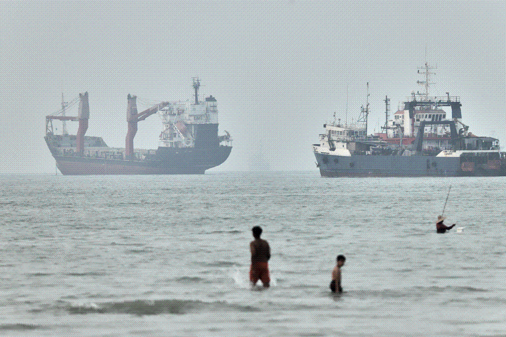 Cargo ships lying at anchor in hazy grey light off a coast in the Strait of Hormuz, with children wading in the shallows and a fisherman in the foreground.