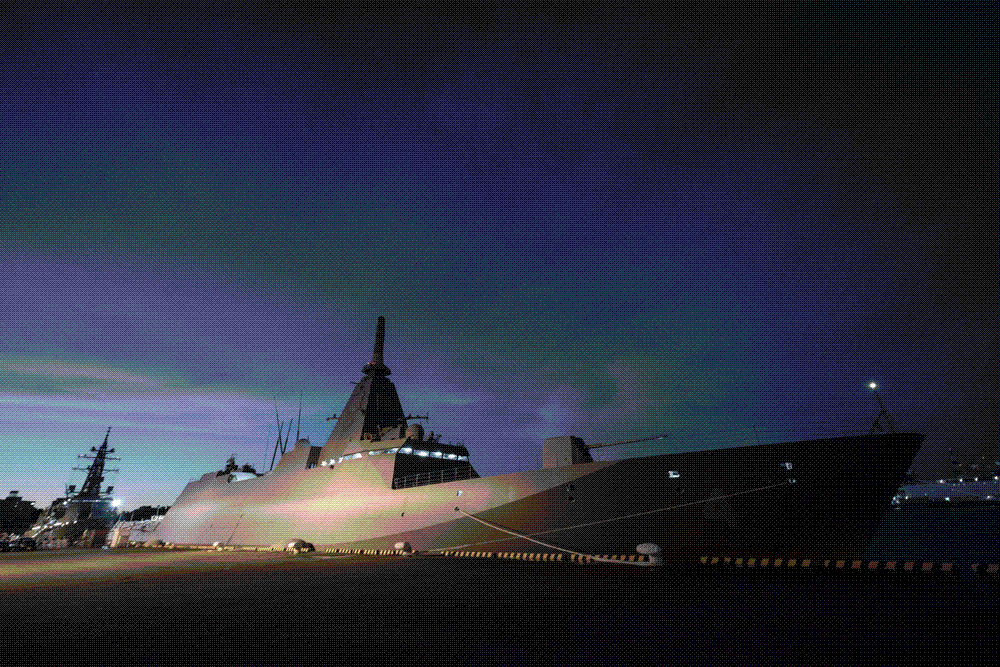 A Japanese naval destroyer moored at a floodlit quayside under a deep twilight sky.