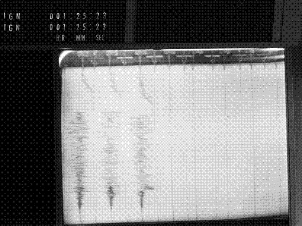 Black-and-white photograph of a seismograph strip chart in a control room: three parallel ink traces run across ruled paper, flat at one end and swelling into thick bands of oscillation at the other, with a digital mission-elapsed-time readout above.