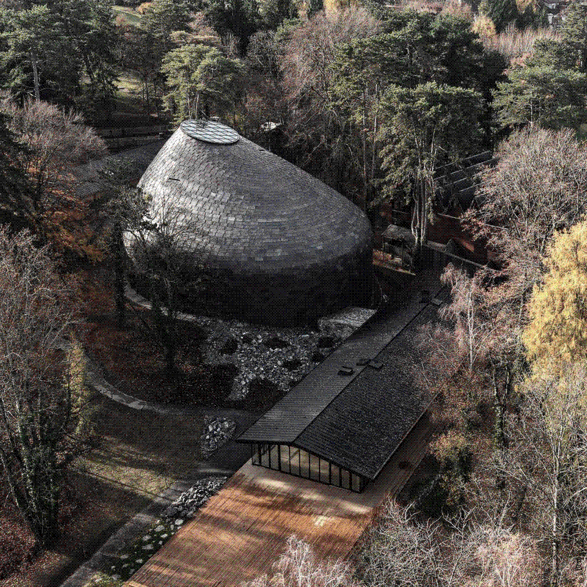 An aerial view of a domed, shingle-clad concert hall nestled among autumn woodland, with a long dark-roofed timber walkway leading to it.