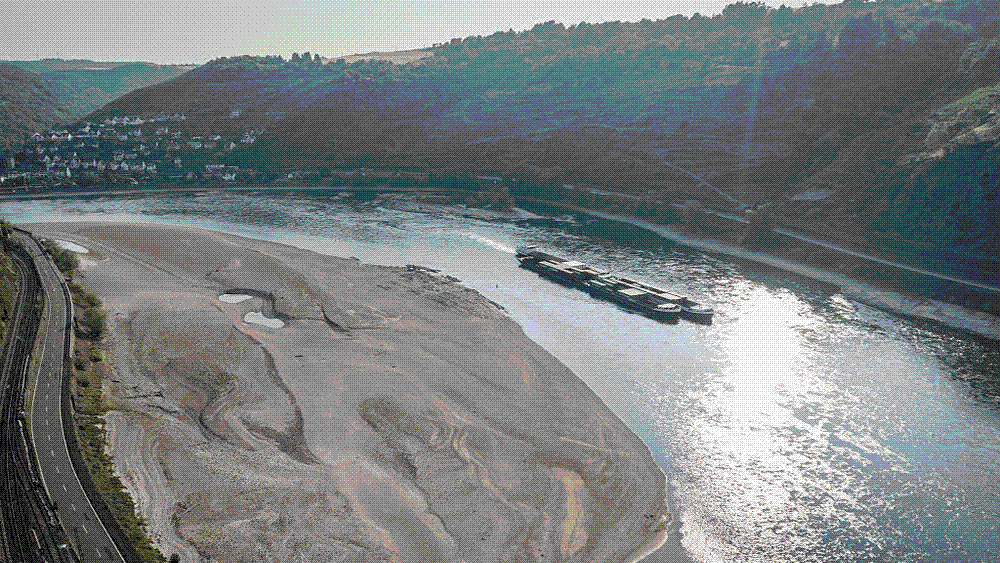 A wide river reduced to a narrow channel between broad expanses of exposed, cracked riverbed and gravel banks under a hazy summer sky.