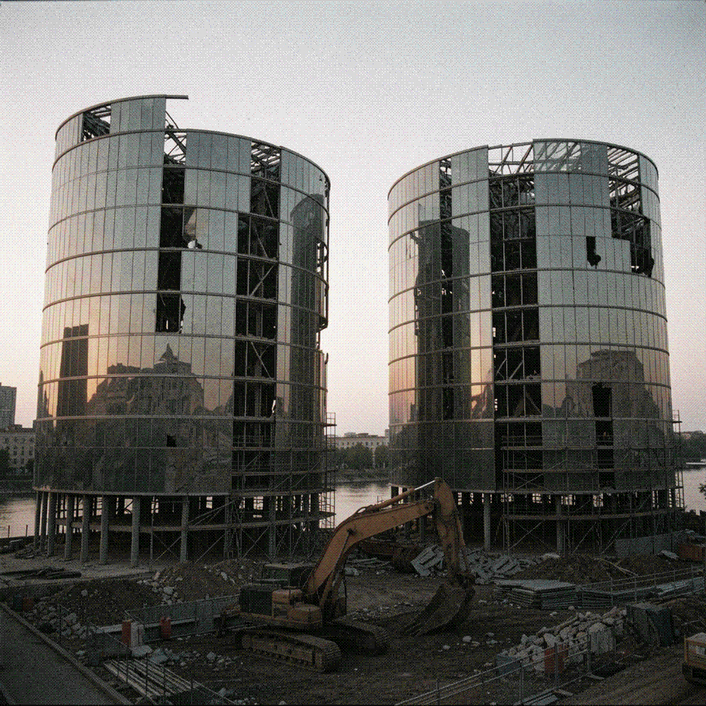 Two tubular glass-and-steel buildings by a river at dusk, curved cladding half stripped to the frame, scaffolding below