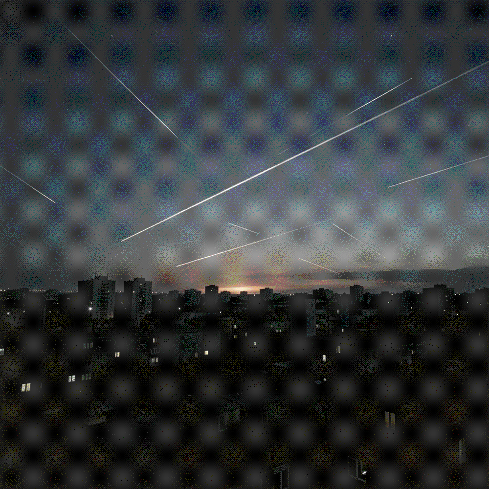 Darkened city skyline at night crossed by thin bright streaks of light, a faint orange glow on the horizon