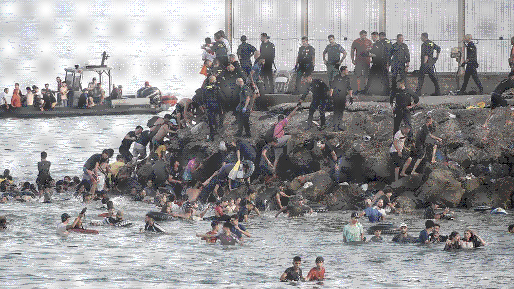 A tall border fence topped with razor wire running down toward a rocky Mediterranean shoreline.