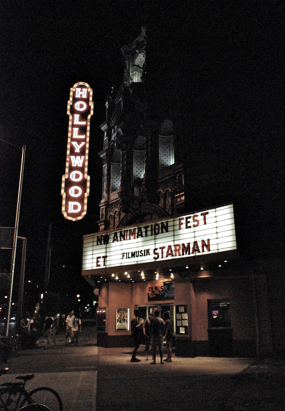A grand movie palace's illuminated marquee and vertical blade sign blaze against the night sky.