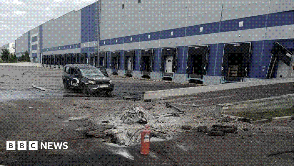 A blast crater and scattered rubble on the apron outside a long modern distribution warehouse, with a damaged car standing beside torn loading-bay doors.